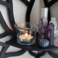 A black wooden wall shelf shaped like a lotus flower, mounted in the corner of a room. The shelf holds a candle in a glass holder, several crystal stones, and a small spray bottle. Decorative and compact, it adds a serene, spiritual touch to the space. Dimensions are 23cm (9.05in) wide and 29cm (11.41in) tall.