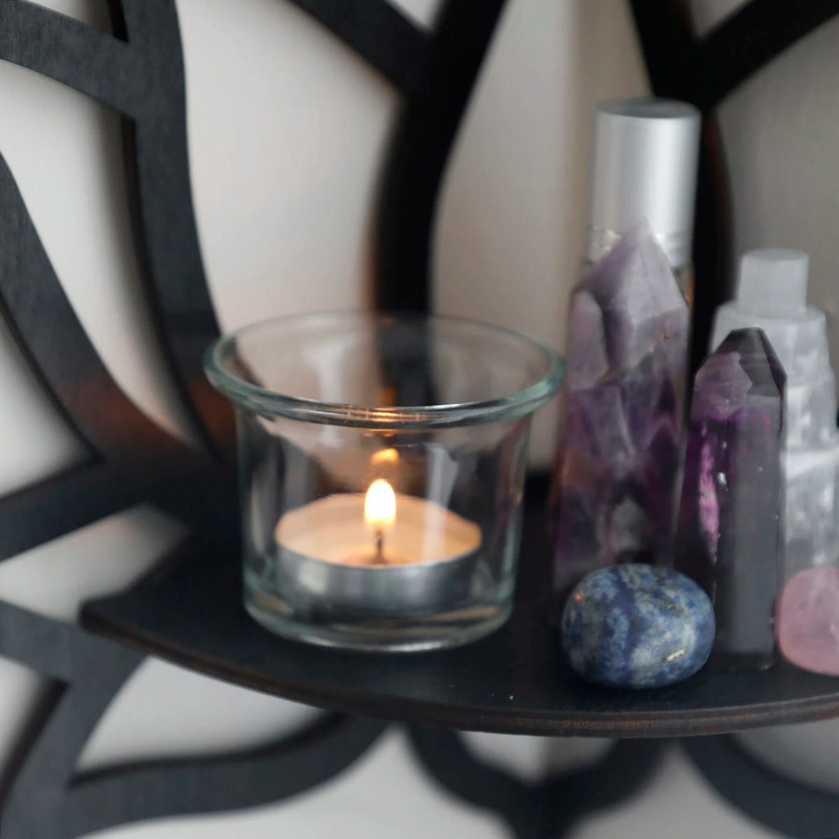 A black wooden wall shelf shaped like a lotus flower, mounted in the corner of a room. The shelf holds a candle in a glass holder, several crystal stones, and a small spray bottle. Decorative and compact, it adds a serene, spiritual touch to the space. Dimensions are 23cm (9.05in) wide and 29cm (11.41in) tall.
