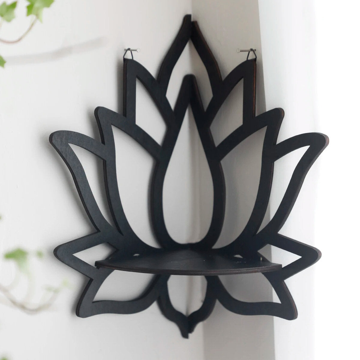 A black wooden wall shelf shaped like a lotus flower, mounted in the corner of a room. The shelf holds a candle in a glass holder, several crystal stones, and a small spray bottle. Decorative and compact, it adds a serene, spiritual touch to the space. Dimensions are 23cm (9.05in) wide and 29cm (11.41in) tall.