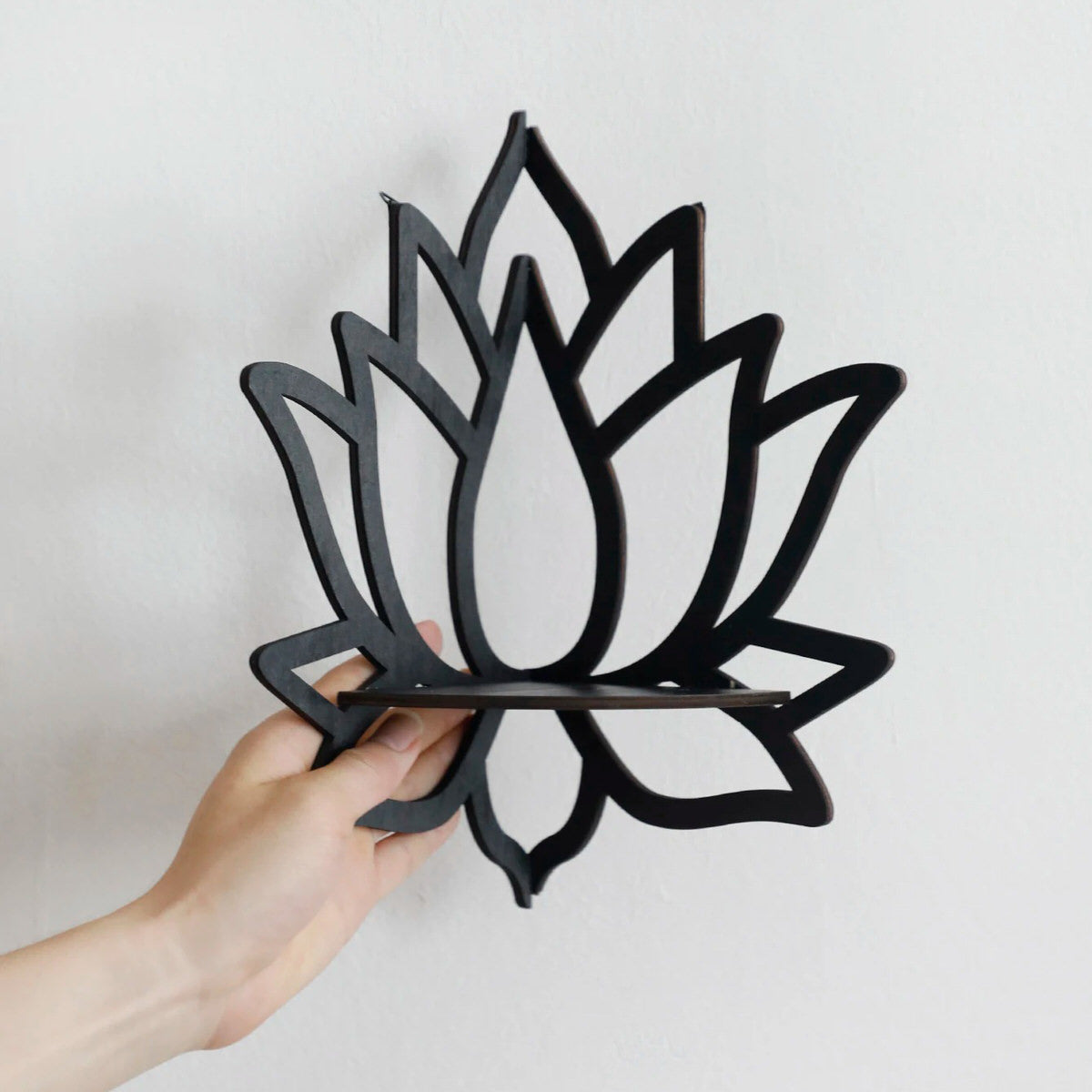 A black wooden wall shelf shaped like a lotus flower, mounted in the corner of a room. The shelf holds a candle in a glass holder, several crystal stones, and a small spray bottle. Decorative and compact, it adds a serene, spiritual touch to the space. Dimensions are 23cm (9.05in) wide and 29cm (11.41in) tall.
