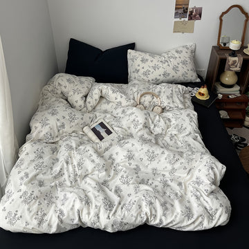 Creamy white bedding set printed with black outlined floral designs, giving a delicate and minimalist look. The bedding is soft and rumpled, styled with black pillows and cozy bedroom decor.