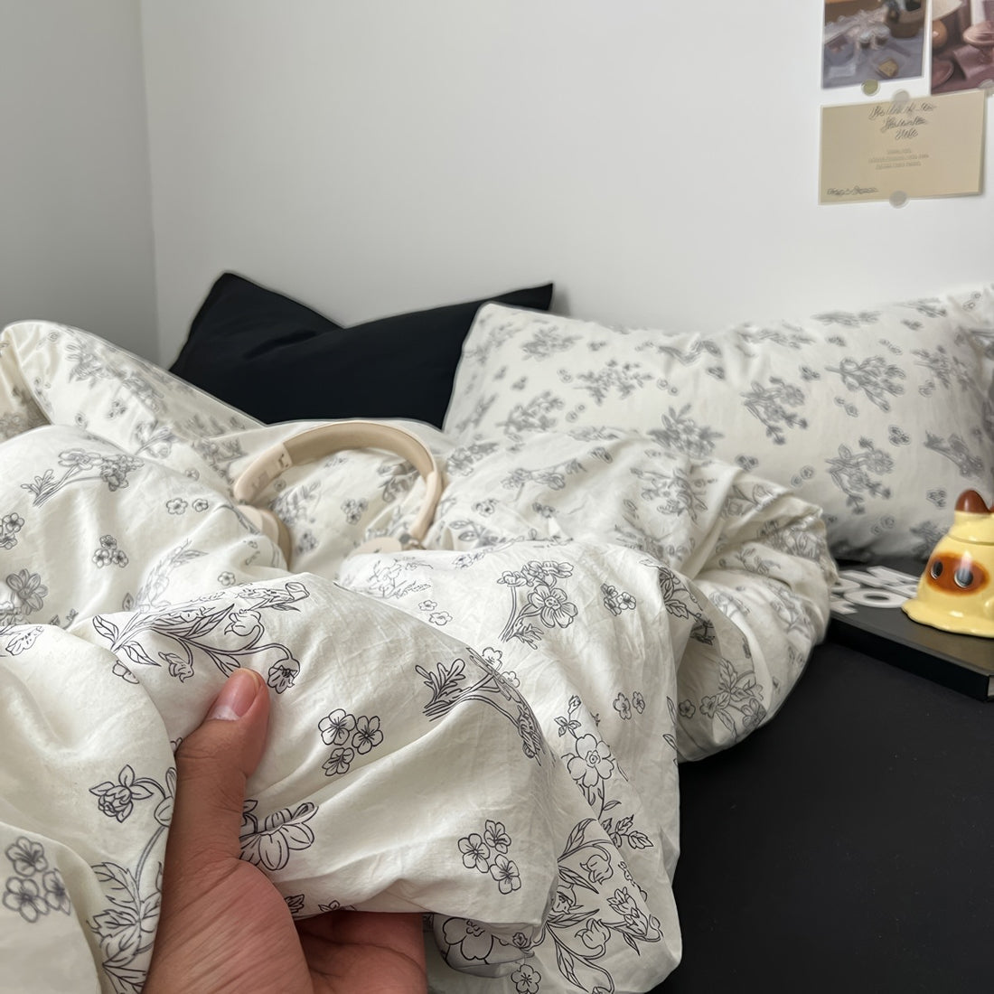 Creamy white bedding set printed with black outlined floral designs, giving a delicate and minimalist look. The bedding is soft and rumpled, styled with black pillows and cozy bedroom decor.
