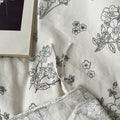 Creamy white bedding set printed with black outlined floral designs, giving a delicate and minimalist look. The bedding is soft and rumpled, styled with black pillows and cozy bedroom decor.