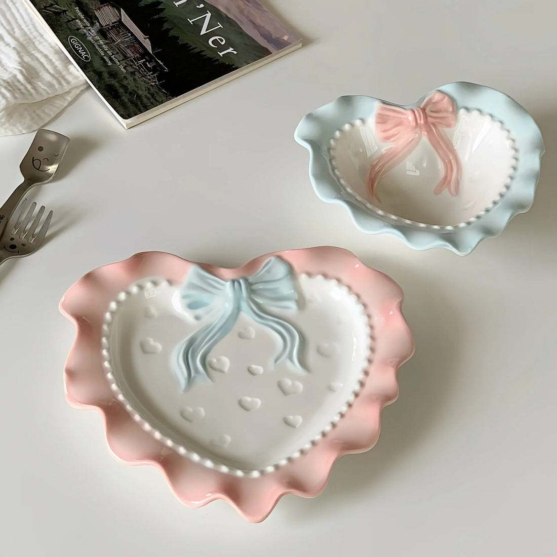 A pastel-colored, heart-shaped ceramic plate featuring scalloped edges, embossed hearts, and an elegant bow accent. Available in soft pink, pastel blue, and delicate yellow colors.