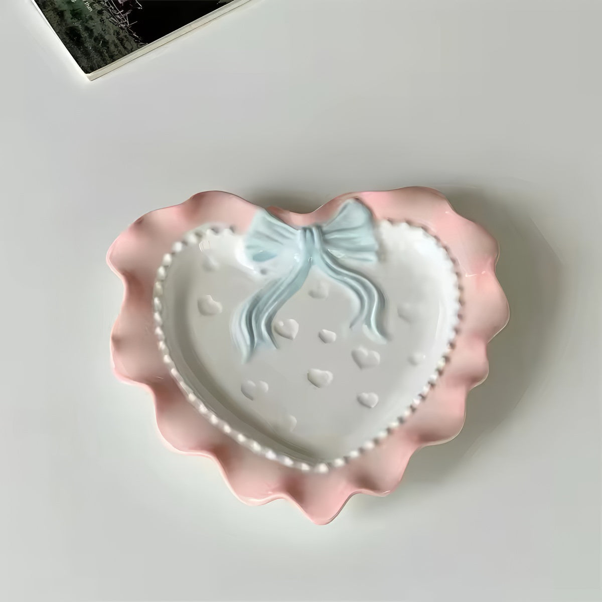 A pastel-colored, heart-shaped ceramic plate featuring scalloped edges, embossed hearts, and an elegant bow accent. Available in soft pink, pastel blue, and delicate yellow colors.