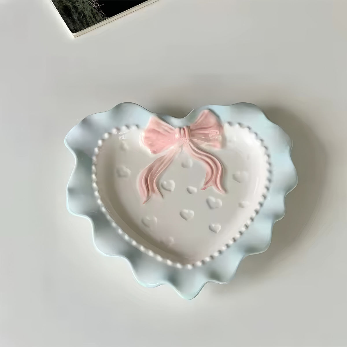 A pastel-colored, heart-shaped ceramic plate featuring scalloped edges, embossed hearts, and an elegant bow accent. Available in soft pink, pastel blue, and delicate yellow colors.