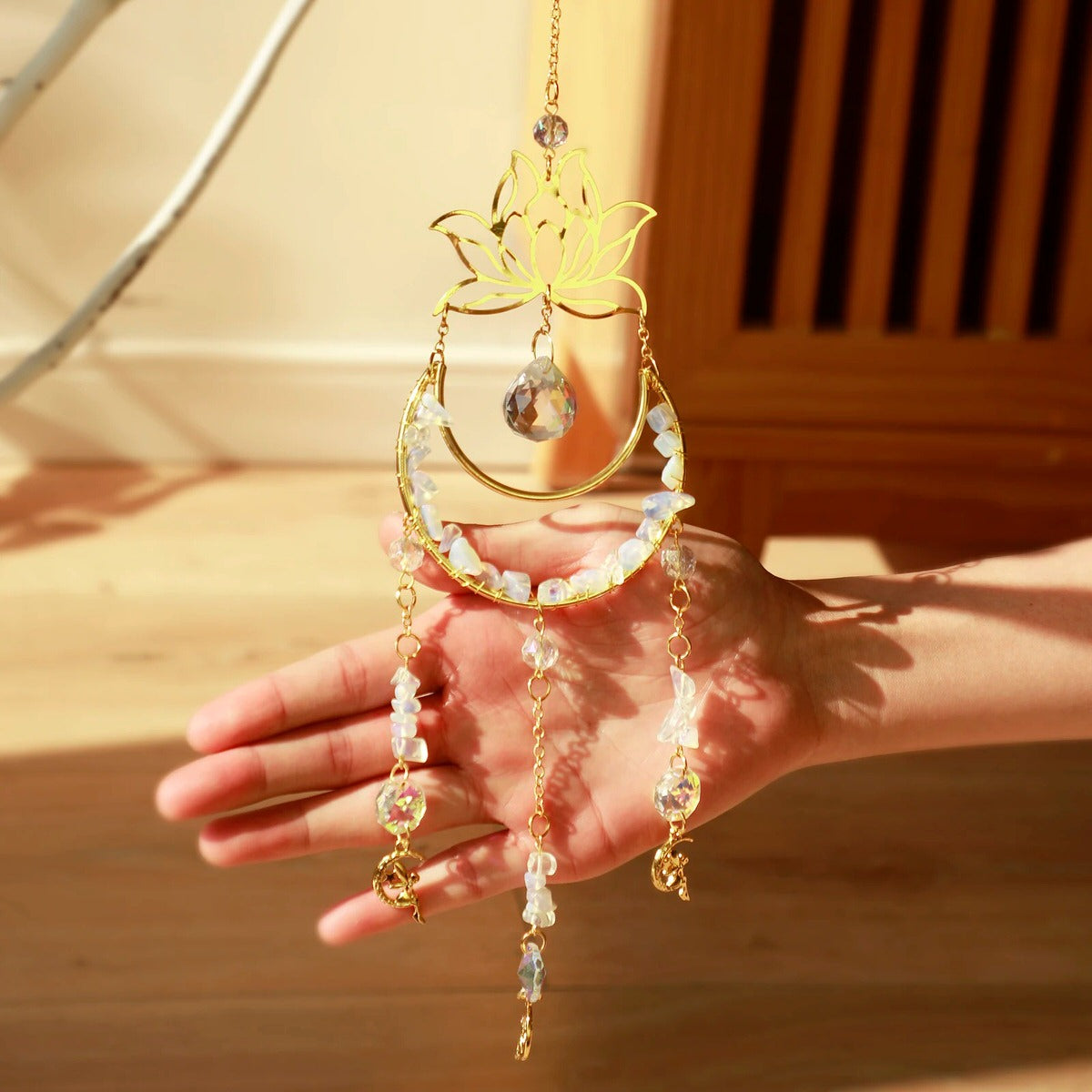A golden suncatcher featuring a lotus flower at the top, a crescent-shaped ring adorned with white crystal chips, and a central hanging prism. Three crystal and charm strands dangle below, reflecting rainbow light patterns onto the surrounding wall. The full hanging length is 44cm (17.32in).