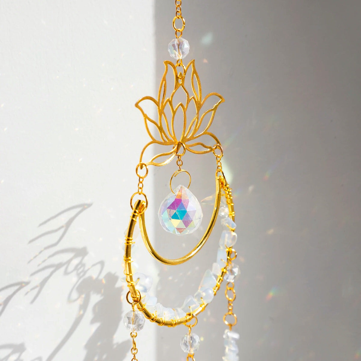 A golden suncatcher featuring a lotus flower at the top, a crescent-shaped ring adorned with white crystal chips, and a central hanging prism. Three crystal and charm strands dangle below, reflecting rainbow light patterns onto the surrounding wall. The full hanging length is 44cm (17.32in).