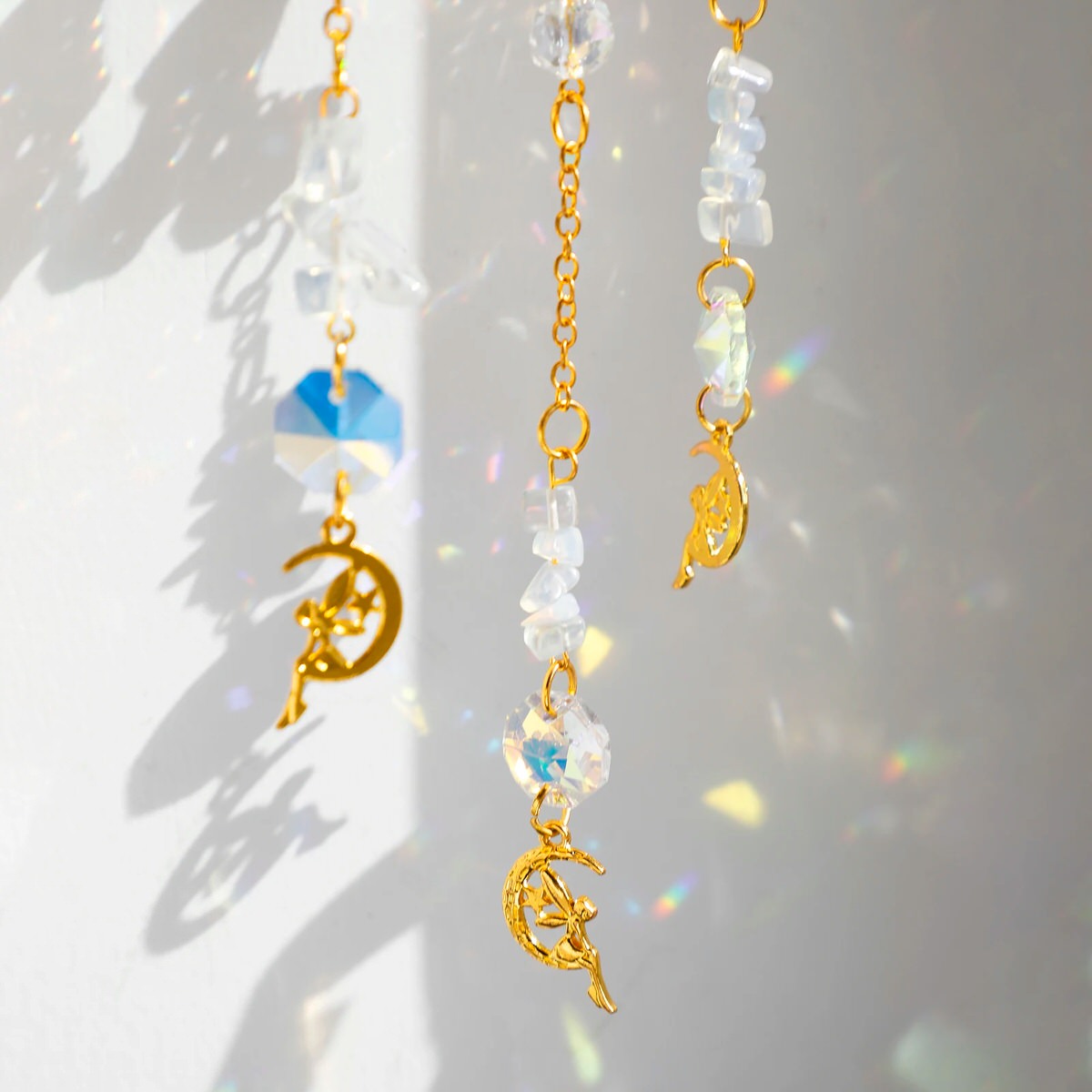A golden suncatcher featuring a lotus flower at the top, a crescent-shaped ring adorned with white crystal chips, and a central hanging prism. Three crystal and charm strands dangle below, reflecting rainbow light patterns onto the surrounding wall. The full hanging length is 44cm (17.32in).