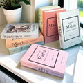 Set of five minimalist faux book storage boxes styled as fashion city books (Paris, Milan, London, Berlin, Madrid) on a window shelf – hidden storage and decorative display.