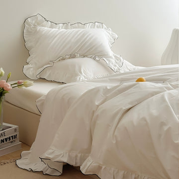 White bedding set with ruffled pillowcases trimmed in black piping, styled in a minimal bedroom.