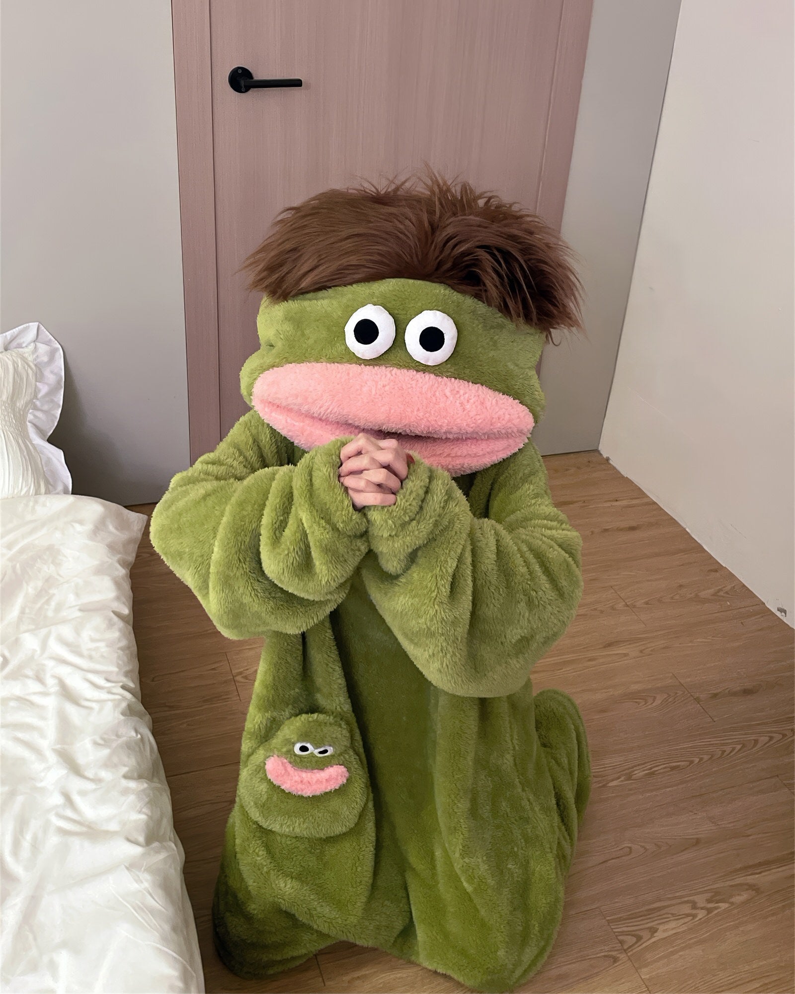 A green plush hooded one-piece pajama or costume with a frog-like face, large white eyes, a pink mouth, and a small matching frog patch on the chest