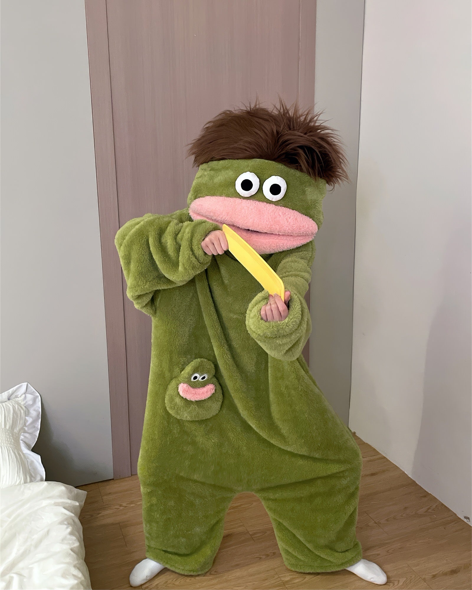 A green plush hooded one-piece pajama or costume with a frog-like face, large white eyes, a pink mouth, and a small matching frog patch on the chest