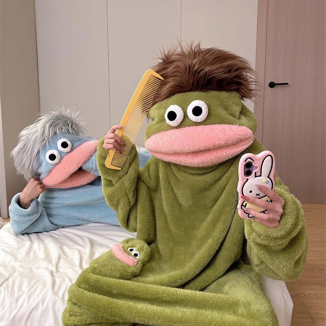 A green plush hooded one-piece pajama or costume with a frog-like face, large white eyes, a pink mouth, and a small matching frog patch on the chest