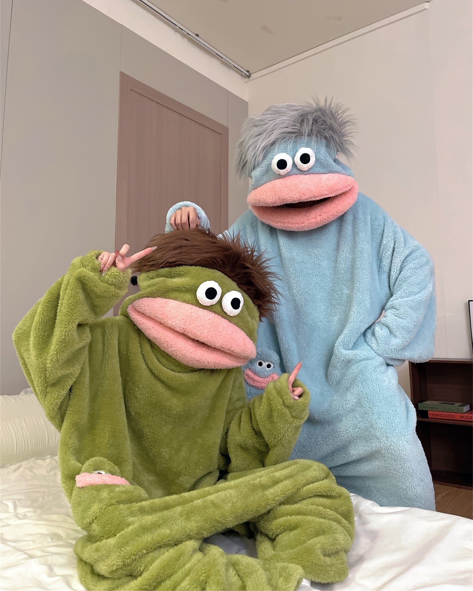 A green plush hooded one-piece pajama or costume with a frog-like face, large white eyes, a pink mouth, and a small matching frog patch on the chest