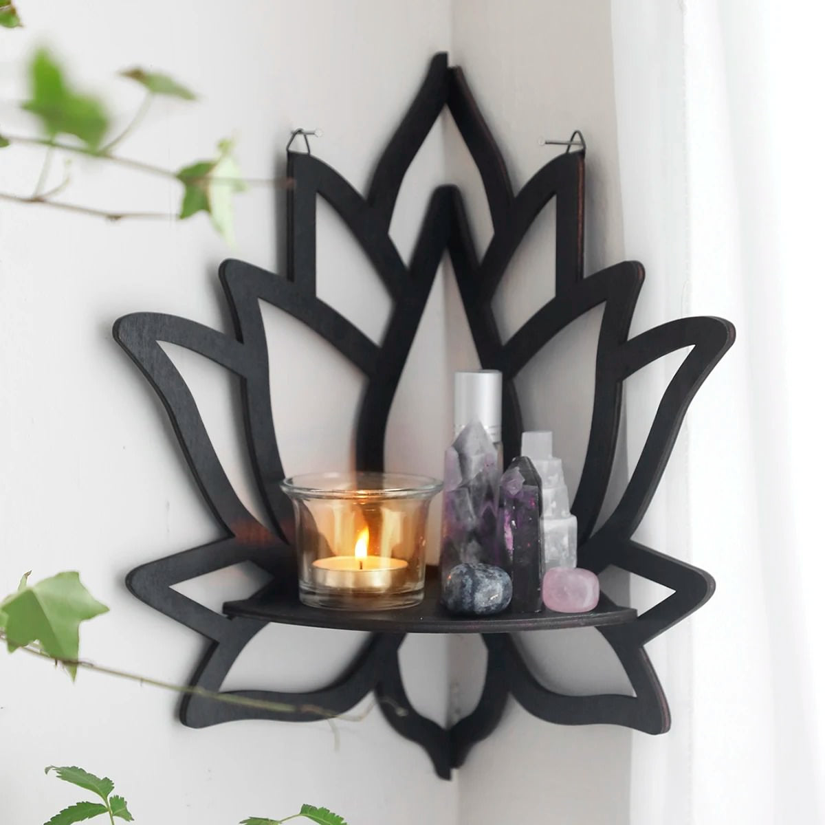 A black wooden wall shelf shaped like a lotus flower, mounted in the corner of a room. The shelf holds a candle in a glass holder, several crystal stones, and a small spray bottle. Decorative and compact, it adds a serene, spiritual touch to the space. Dimensions are 23cm (9.05in) wide and 29cm (11.41in) tall.