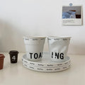 White ceramic coffee mug shaped like a crumpled takeaway coffee cup with black TOASTING lettering and #coffee print around the rim.