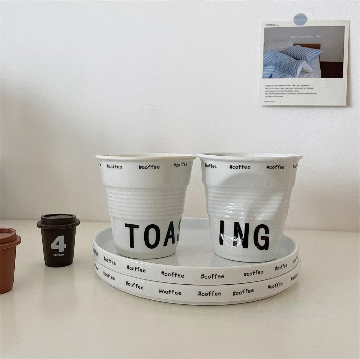 White ceramic coffee mug shaped like a crumpled takeaway coffee cup with black TOASTING lettering and #coffee print around the rim.