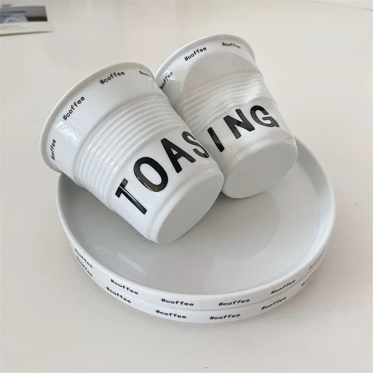 White ceramic coffee mug shaped like a crumpled takeaway coffee cup with black TOASTING lettering and #coffee print around the rim.