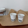 White ceramic coffee mug shaped like a crumpled takeaway coffee cup with black TOASTING lettering and #coffee print around the rim.