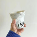 White ceramic coffee mug shaped like a crumpled takeaway coffee cup with black TOASTING lettering and #coffee print around the rim.