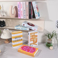 Decorative faux book storage box designed as a colorful hardcover book with a hidden storage compartment for organizing small household items while decorating shelves and tables