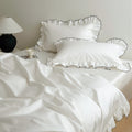 White bedding set with ruffled pillowcases trimmed in black piping, styled in a minimal bedroom.