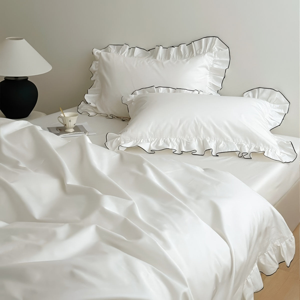 White bedding set with ruffled pillowcases trimmed in black piping, styled in a minimal bedroom.