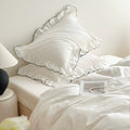 White bedding set with ruffled pillowcases trimmed in black piping, styled in a minimal bedroom.